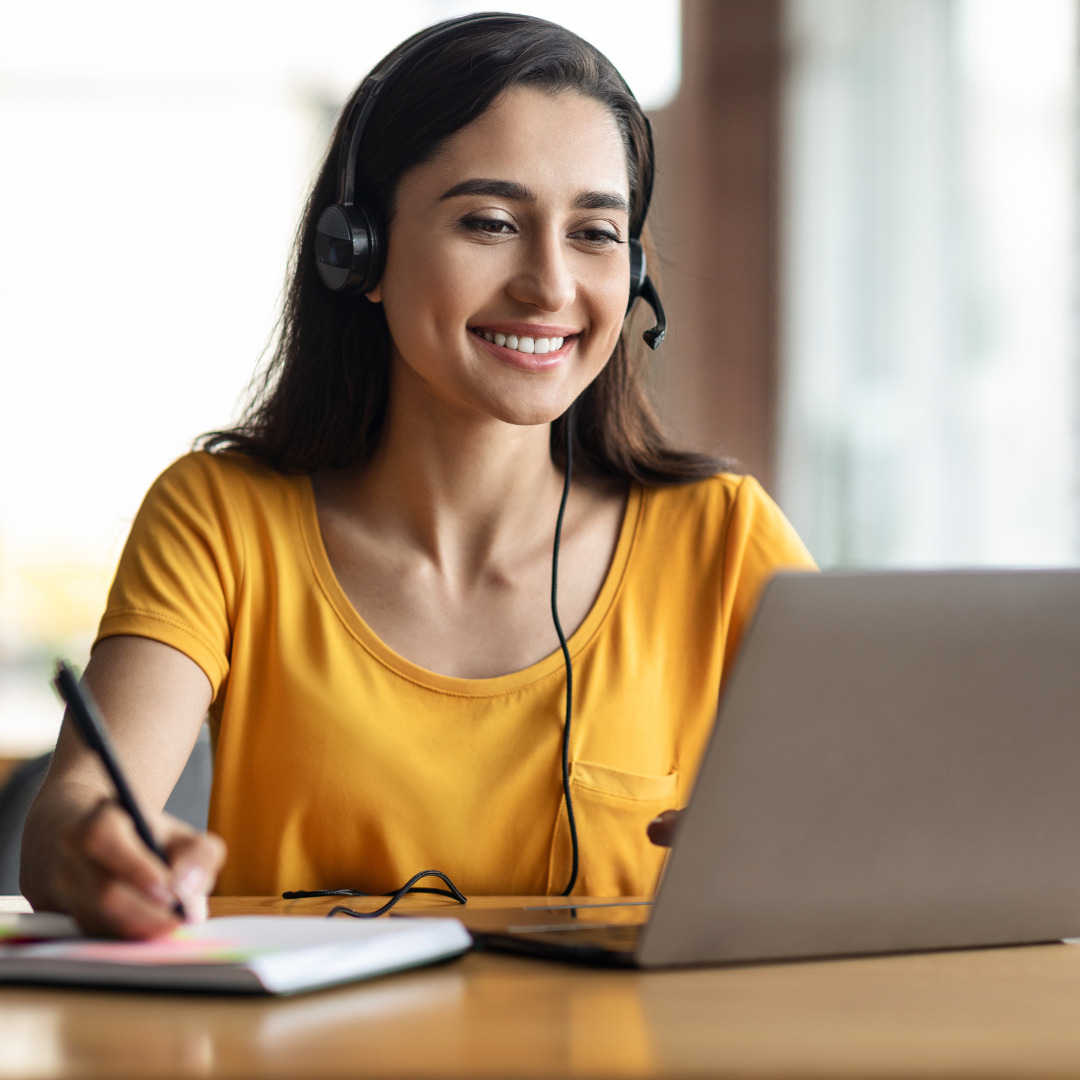 A person with a headset on taking notes while listening to a class on a laptop