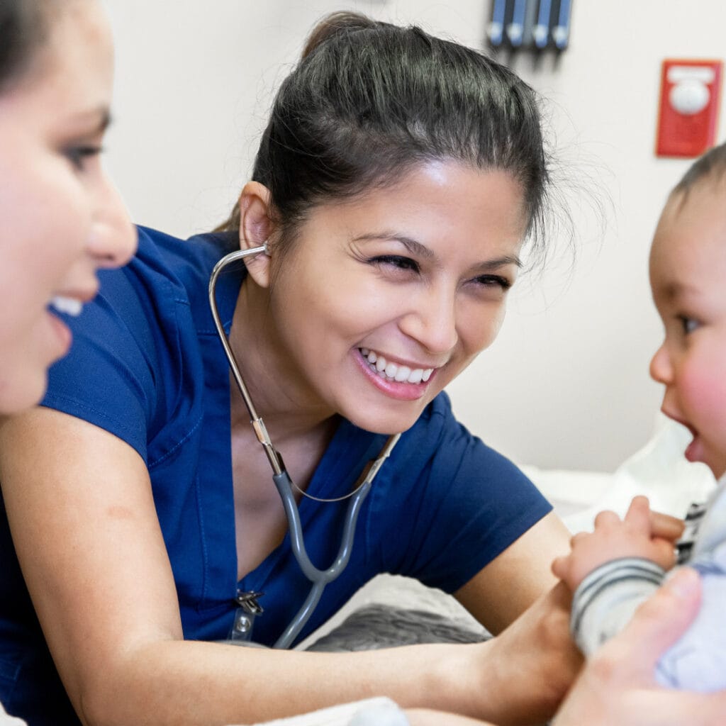 A smiling nurse practitioner listening to a baby's heartbeat as the baby's mother looks on